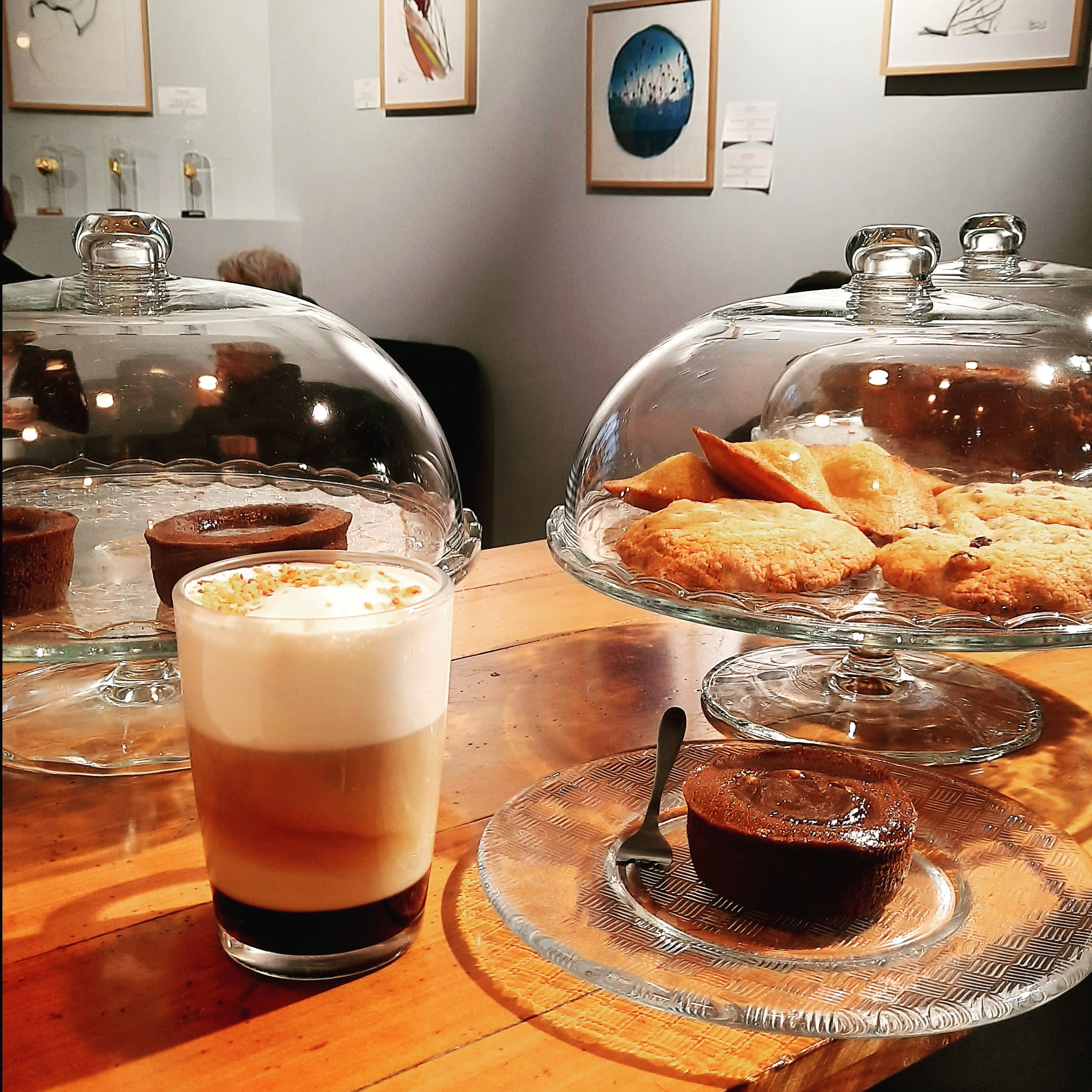 Photo d'un latte macchiato avec un moelleux au chocolat à côté. Derrière des cloches en verre couvrant des pâtisseries. Au fond la galerie d'art de L'Equilibre du monde, café salon de thé librairie,à Saint-Renan Pays d'Iroise Brest